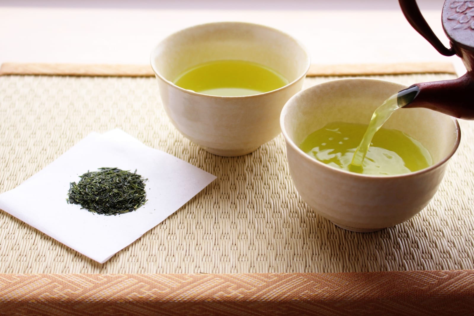 Sencha poured from a clay teapot into two small cups, with loose sencha leaves beside them